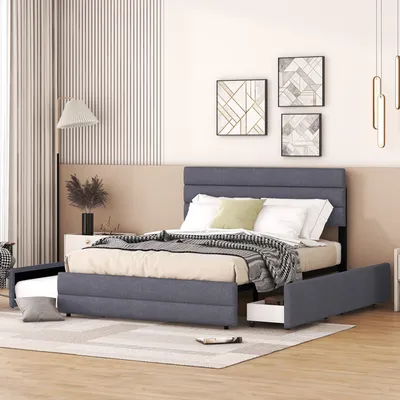 A bedroom scene featuring a gray upholstered bed with a pull-out trundle bed and drawers, decorated with artwork and a floor lamp.