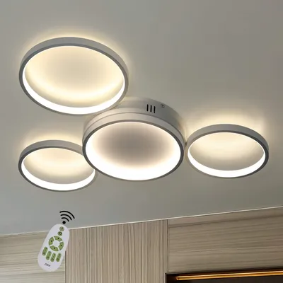 A modern ceiling light fixture with four circular rings of varying sizes, illuminated and mounted on a white ceiling. A remote control is visible in the lower left corner.