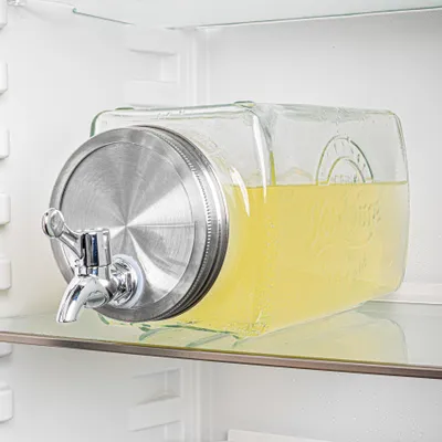 A glass beverage dispenser filled with yellow liquid, placed on a shelf inside a refrigerator.