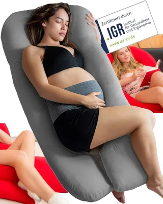 A pregnant woman is sleeping on a large gray body pillow, while another woman reads a book in the background.