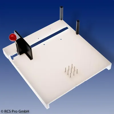 A white cutting board with various features, including a red-handled lever, a slot, and metal pins, is shown against a blue background.