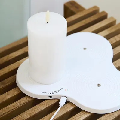 A white LED candle sits on a white charging base, which is placed on a wooden slatted surface.