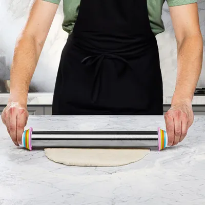 A person wearing a black apron is using a stainless steel rolling pin with colorful adjustable thickness rings to roll out dough on a marble countertop.