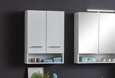 Two white bathroom cabinets are shown against a dark gray wall. The cabinet on the left has two doors and an open shelf below, containing towels and containers. The cabinet on the right has mirrored doors and an open shelf below, holding decorative items a