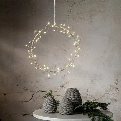 A circular LED wreath with warm white lights hangs against a textured, light gray wall. Below, on a round table, are three decorative pinecone-shaped objects and some greenery.