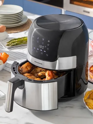 A black and silver air fryer with chicken wings inside, placed on a kitchen counter with other food items and plates in the background.