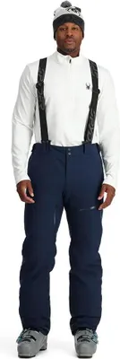 A man wearing ski gear, including a white jacket, navy ski pants, and a beanie, stands against a white background.
