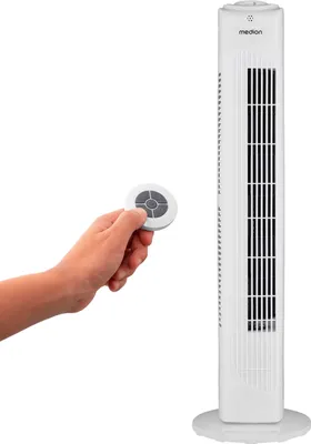 A hand holds a remote control in front of a tall white tower fan.