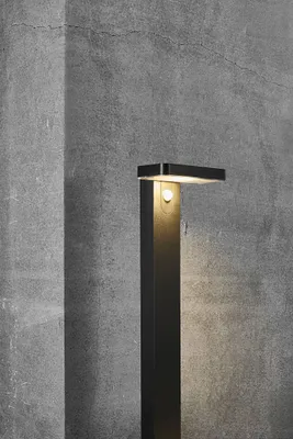 A modern black LED garden light with a solar panel, set against a textured gray concrete wall.