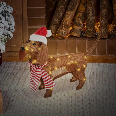 A lighted dachshund decoration with a Santa hat and scarf sits in front of a fireplace.