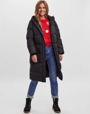 A woman is standing and posing in a studio setting, wearing a long black puffer jacket, a red sweater with white snowflake designs, and blue jeans.