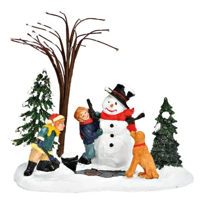 Zwei Kinder die dabei sind einen Schneemann zu bauen, und ihr kleiner Hund sitzt ganz brav daneben.