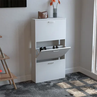 A white shoe cabinet with three flip-down drawers, one of which is open showing shoes inside. The cabinet is placed against a light gray wall, with a wooden shelf unit to the left and a vase with flowers on top of the cabinet.