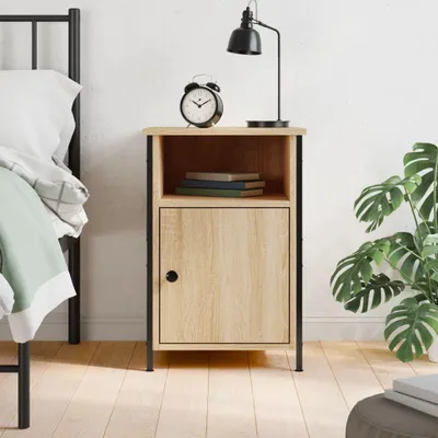 A bedside table with a lamp, alarm clock, and books on top, next to a bed and a plant.