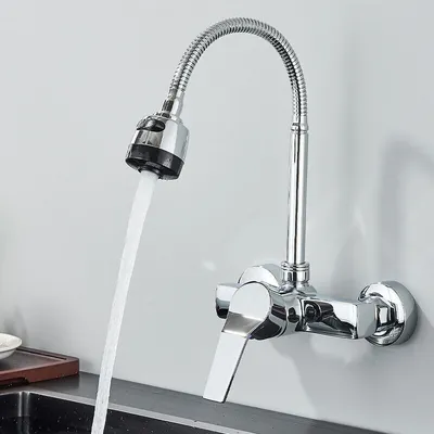 A chrome faucet with a flexible hose and spray head is mounted on a wall, with water flowing into a sink.