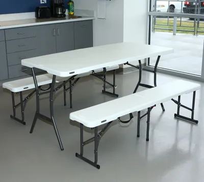 A white plastic folding table and two benches are set up in a room with gray cabinets and a large window overlooking an outdoor area.