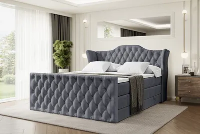 A luxurious dark gray velvet box spring bed with a tufted headboard and footboard, set in a bright, modern bedroom with large windows and a nightstand.