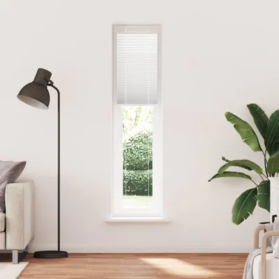 A tall, narrow window with white blinds is shown in the center of a white wall. To the left, a floor lamp and part of a sofa are visible. To the right, a large potted plant with broad green leaves is present. The floor is made of wooden planks.