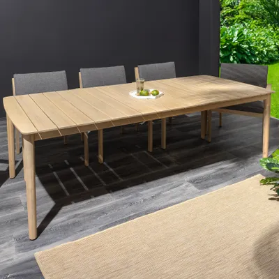 An outdoor patio setting with a wooden dining table and chairs, a rug, and greenery in the background.