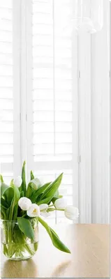 A vase of white tulips sits on a wooden table in front of white shutters.