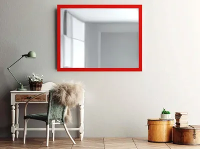 A mirror with a red frame hangs on a wall above a desk with a lamp and a chair.