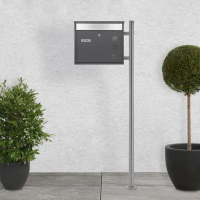 A mailbox with a newspaper compartment mounted on a stand, placed outdoors next to a wall and potted plants.