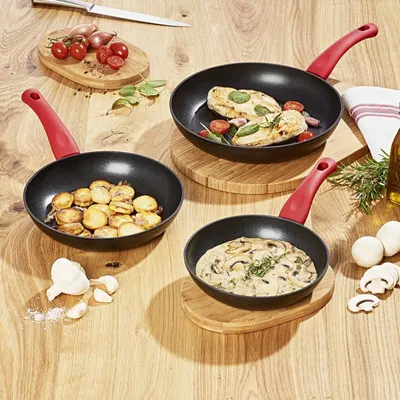 Three frying pans with food being cooked in them, arranged on a wooden table with ingredients like shallots, tomatoes, garlic, and mushrooms.