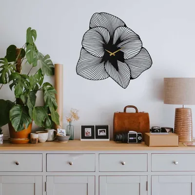 A black metal wall clock shaped like a flower is mounted on a white wall above a wooden dresser. The dresser is decorated with various items, including plants, picture frames, a vintage suitcase, and a lamp.