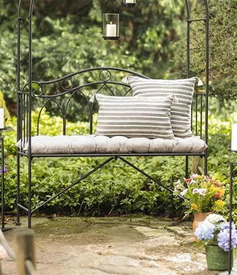 A garden bench with cushions and pillows, surrounded by greenery and flowers.