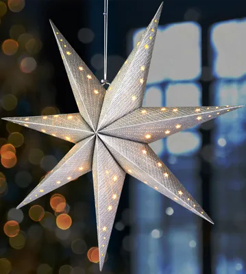 A silver star-shaped lantern with small yellow lights is hanging in front of a blurred background of lights and a window.