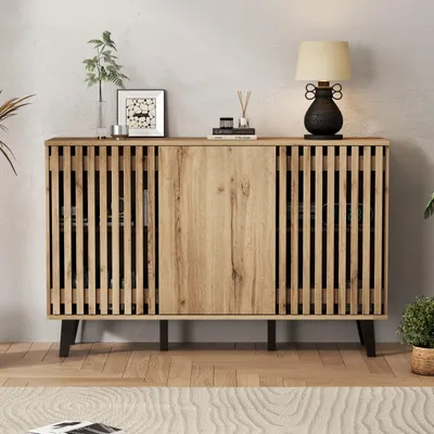A wooden sideboard with slatted doors and black legs, decorated with a lamp, a framed picture, and a vase with a plant.