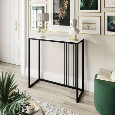 A console table with a white top and black metal frame, decorated with vertical slats on one side, is placed in a well-lit room with framed artwork on the wall and a green ottoman nearby.