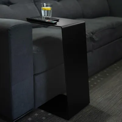 A black C-shaped side table is placed next to a dark gray sofa, with a glass of water and a remote control on top.