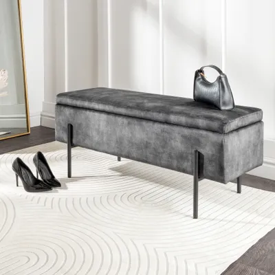 A gray velvet storage bench with black metal legs, a black handbag, and black high heels on a white rug.