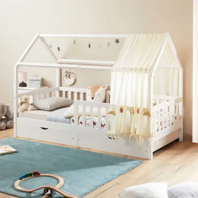A white house-shaped bed for children, with a canopy and bedding, placed on a blue rug in a room.