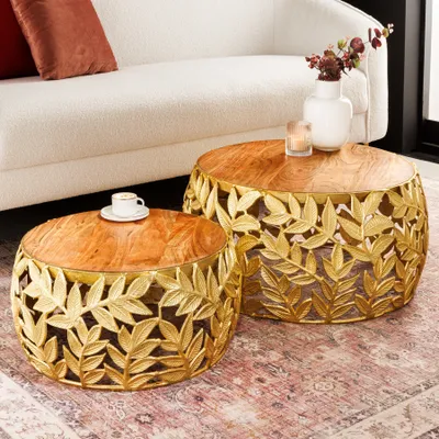 Two round coffee tables with intricate gold leaf designs on their bases and wooden tops, placed on a patterned rug next to a white sofa with a red cushion.