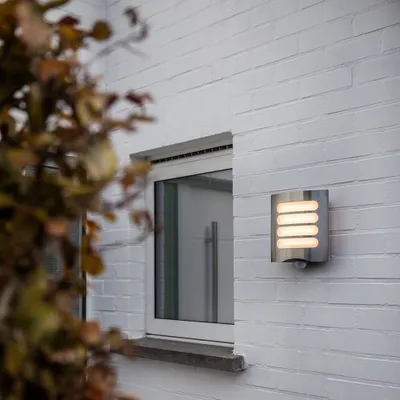 An outdoor wall light is mounted on a white brick wall next to a window. Some autumn foliage is in the foreground.