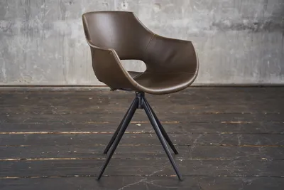 A brown leather chair with a black metal base is shown in a studio setting with a concrete wall and wooden floor.