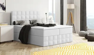 A white boxspring bed with storage is shown in a modern bedroom setting with a yellow rug and a floor lamp.