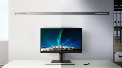 A computer monitor displaying a vibrant aurora borealis scene is placed on a desk. The background transitions from a brightly lit wall on the left to a darker, more shadowed wall on the right, illustrating different lighting environments. A shelf with bind
