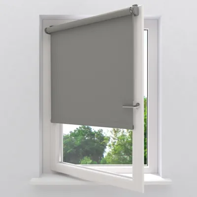 A gray roller blind is installed on a white window frame. The window is open, revealing trees and greenery outside.