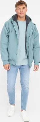 A man wearing a teal jacket and light blue jeans, standing against a white background.