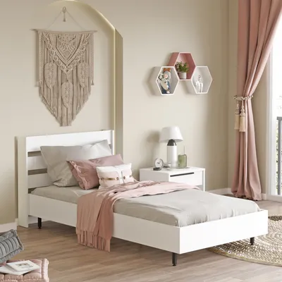 A bedroom scene featuring a white single bed with pink and beige bedding, a macrame wall hanging, and hexagonal shelves.