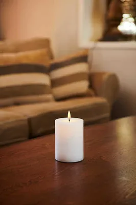 A white LED candle with a lit flame sits on a wooden table in front of a blurred couch.