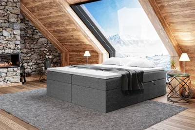 A bedroom scene in a rustic attic with a large window overlooking a snowy mountain landscape. The bed is the central focus, with a fireplace on the left and a side table on the right.
