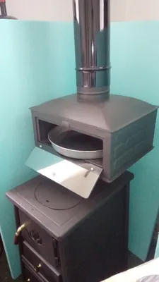 A black wood-burning stove with a pizza oven attachment and a metal chimney pipe, set against a light blue wall.