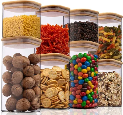 Set aus klaren quadratischen Vorratsbehältern mit Bambusdeckeln, gefüllt mit verschiedenen Trockenwaren wie Nudeln, Kaffeebohnen, Walnüssen, Crackern und Süßigkeiten