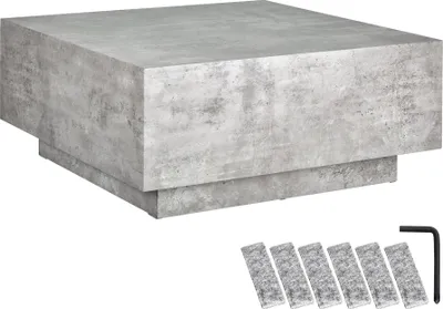 A square coffee table with a concrete-like finish is shown from a diagonal angle. Accessories, including an Allen wrench and several small rectangular pads, are placed in the foreground.