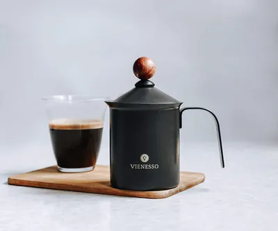A black milk frother with a wooden knob on top sits next to a glass of coffee on a wooden board.
