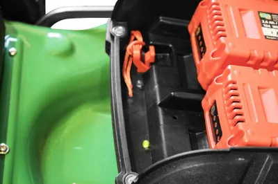 A close-up view of the battery compartment of a green lawnmower, showing two orange batteries and an orange tie-down strap.